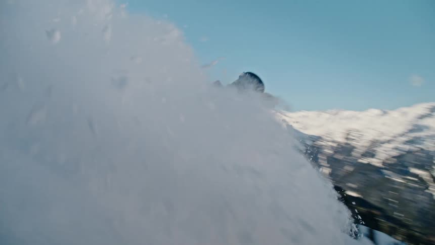 A snowboarder carves down a steep mountain slope, kicking up a massive spray of fresh powder snow that obscures the view in slow motion