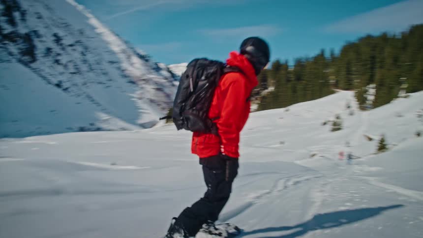 Following a snowboarder in a bright red jacket as they glide through a beautiful snowy mountain landscape on a sunny day