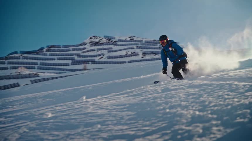 A man in a blue jacket expertly skis down a snowy mountain slope in slow motion on a bright, sunny day