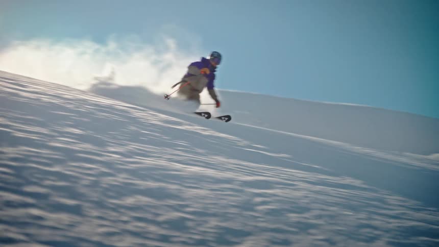 An expert skier carves down a snowy mountain slope in slow motion, kicking up a huge cloud of fresh powder into the bright sun