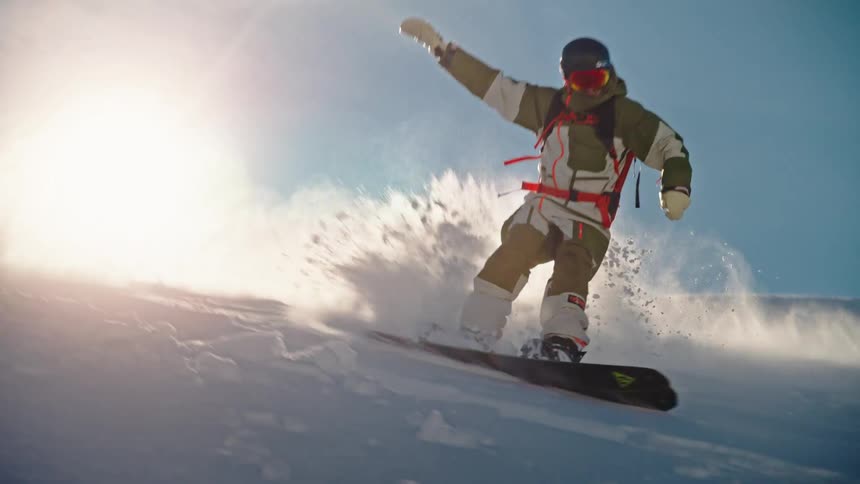 A snowboarder carves down a pristine snowy slope in slow motion, kicking up powder snow against the bright sun
