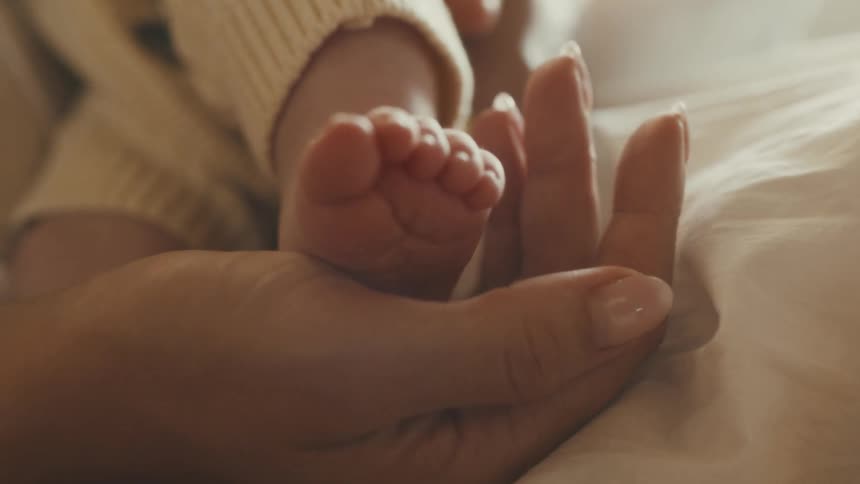 A mother gently holds her newborn baby's tiny foot in her hand as the baby's toes wiggle