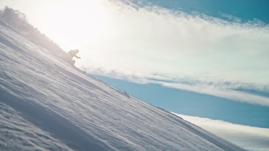 A skier carves down a steep, sun-drenched mountain slope, kicking up fresh powder in slow motion