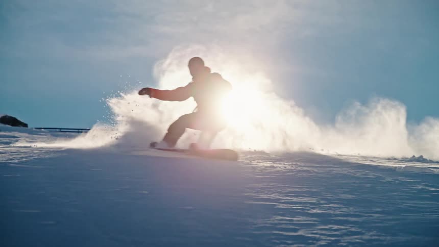 A snowboarder carves through fresh powder snow down a mountain in slow motion, silhouetted against the bright sun