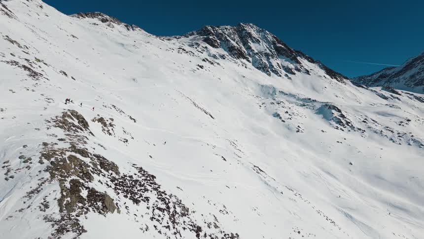 An aerial view follows a group of backcountry skiers as they traverse a snowy mountain ridge before one skis down into fresh powder