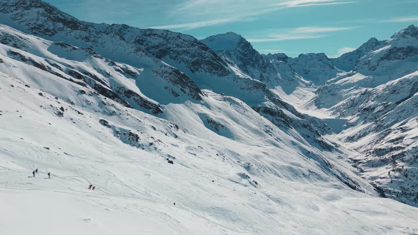 A wide scenic shot shows a group of skiers making their way down a vast, snow-covered mountain valley on a bright winter day