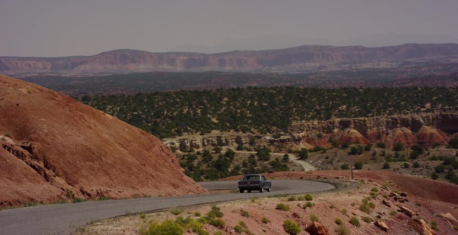A vintage muscle car drives along a winding, empty road through a vast and arid red rock desert landscape