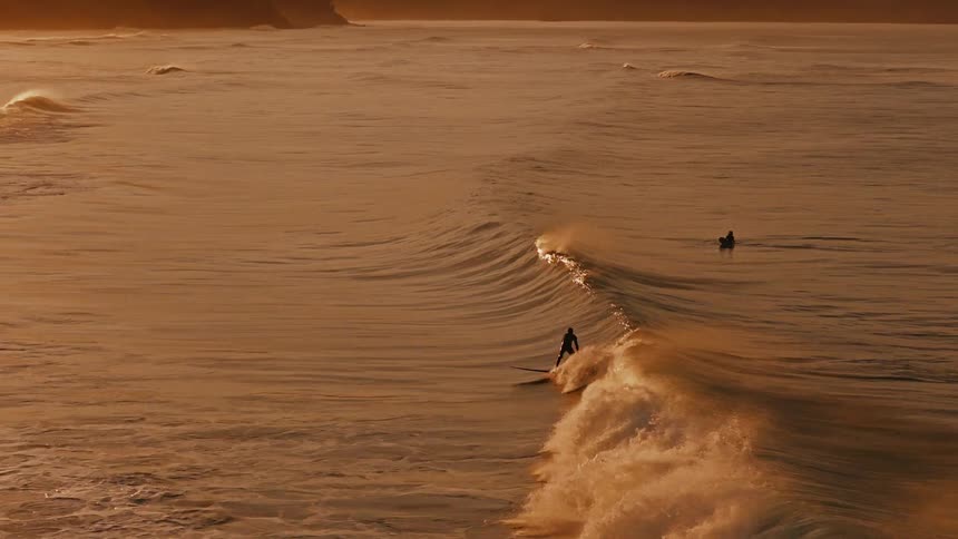 A lone surfer catches and rides a perfect wave in super slow motion against the beautiful golden light of sunset