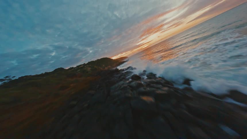 A dynamic FPV drone flies low and fast along a rugged, rocky coastline as waves crash against the shore during a beautiful sunset