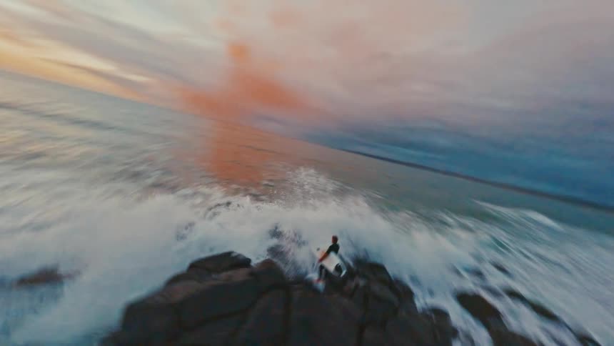 A dynamic FPV drone shot follows a surfer as they jump off a rocky coastline into the ocean waves at sunrise