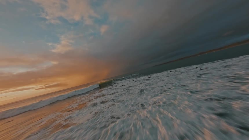 A fast-paced FPV drone shot follows a surfer catching a wave during a dramatic and colorful sunset