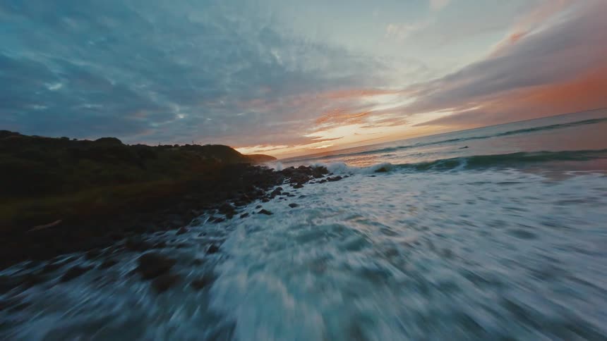 An exhilarating FPV drone speeds low over crashing ocean waves along a rocky coast during a beautiful sunset