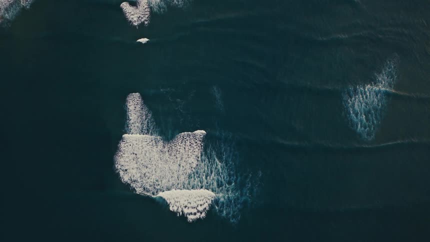 Slow-moving white waves crest over the dark surface of the ocean in this abstract top-down aerial view