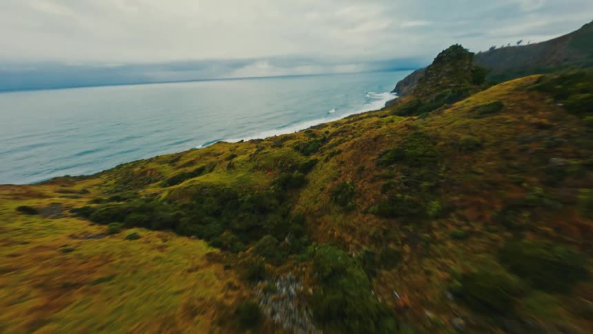 Dynamic FPV drone footage soars over a rugged coastal mountain ridge next to the ocean on a cloudy day
