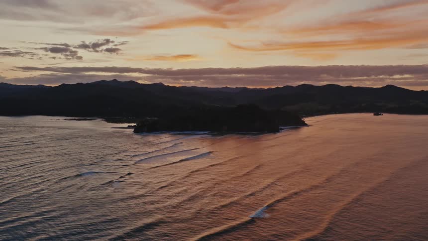 An aerial cinemagraph captures the tranquil beauty of rolling ocean waves reflecting a colorful sunset against a silhouetted coastline