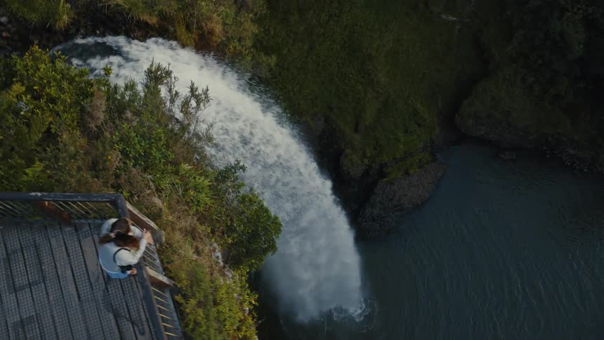 A high-angle aerial view follows a powerful waterfall as a woman watches from a scenic viewing platform surrounded by lush greenery