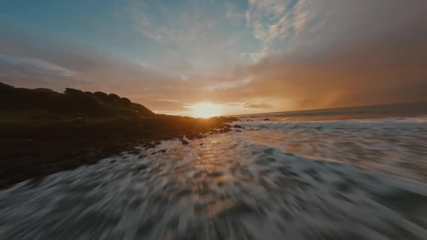 A fast-paced FPV drone flight low over crashing ocean waves along a rocky coastline during a beautiful golden sunset