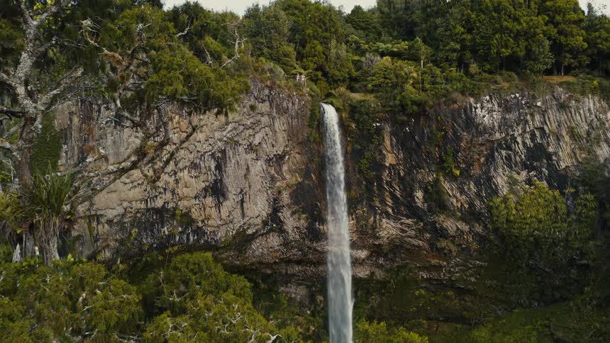 A cinematic aerial shot slowly pushes towards a beautiful waterfall cascading down a sheer rock cliff in a lush green jungle