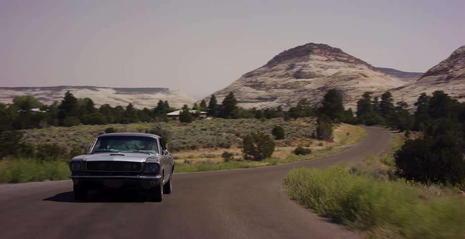 A classic silver Ford Mustang cruises down a winding desert road surrounded by scenic white rock formations