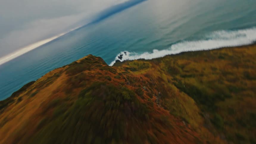 An FPV drone speeds along a windswept cliff before dramatically diving down its rugged face towards the ocean below