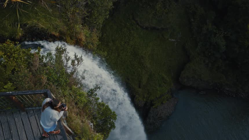 A high-angle aerial shot reveals a woman on a scenic viewpoint photographing a powerful waterfall in a lush forest