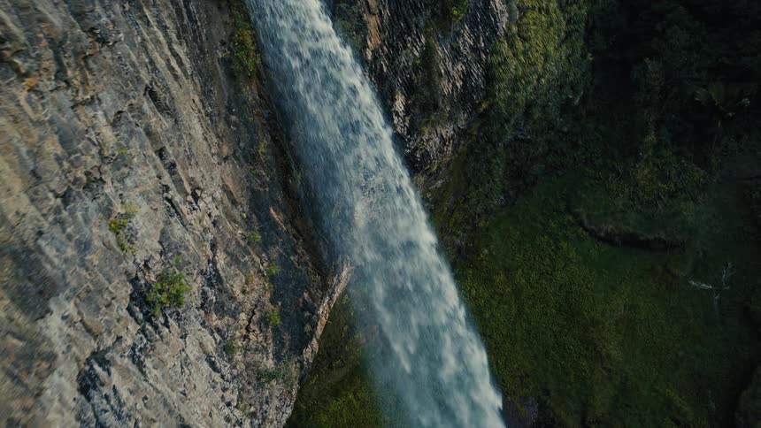 A vertical, close-up aerial shot follows a waterfall as it pours down a rugged, moss-covered cliff face