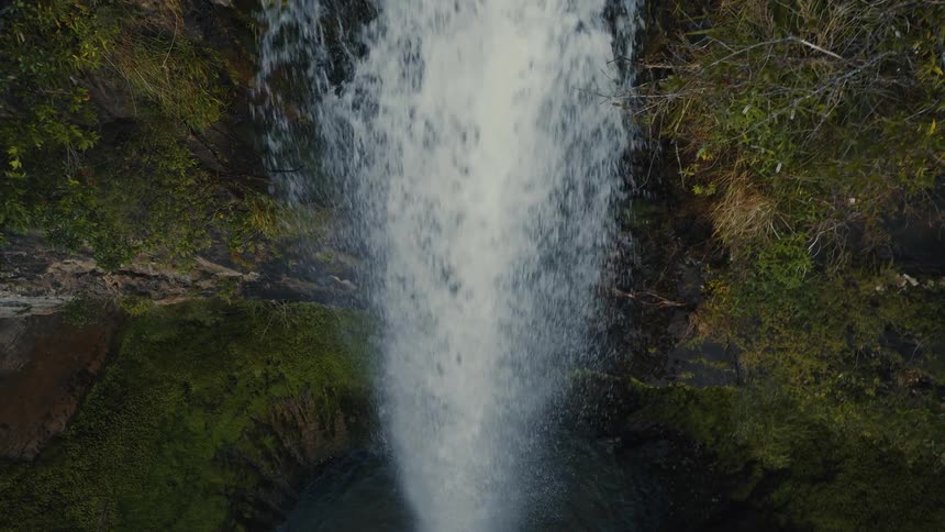 An aerial top-down view of a powerful waterfall cascading down a mossy cliff into a pool below