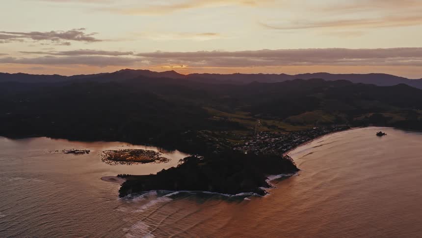 A stunning aerial view captures a coastal town on a peninsula as the sun sets behind the silhouetted mountains