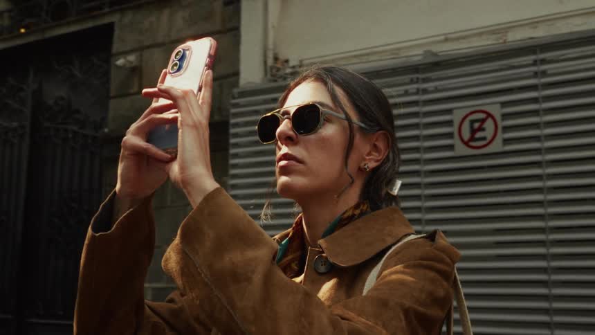 A stylish young woman in sunglasses captures a photo with her smartphone on a city street