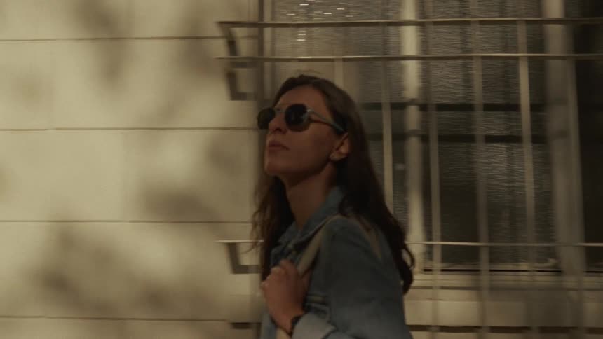 A young woman wearing sunglasses and a jean jacket walks along a city street in the warm afternoon sun