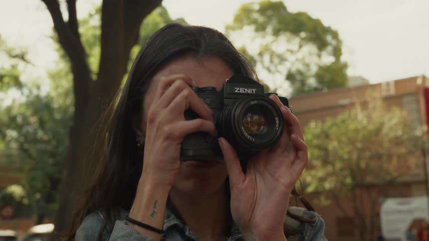 A young woman in a denim jacket points a vintage Zenit camera directly at the viewer on a sunny city street