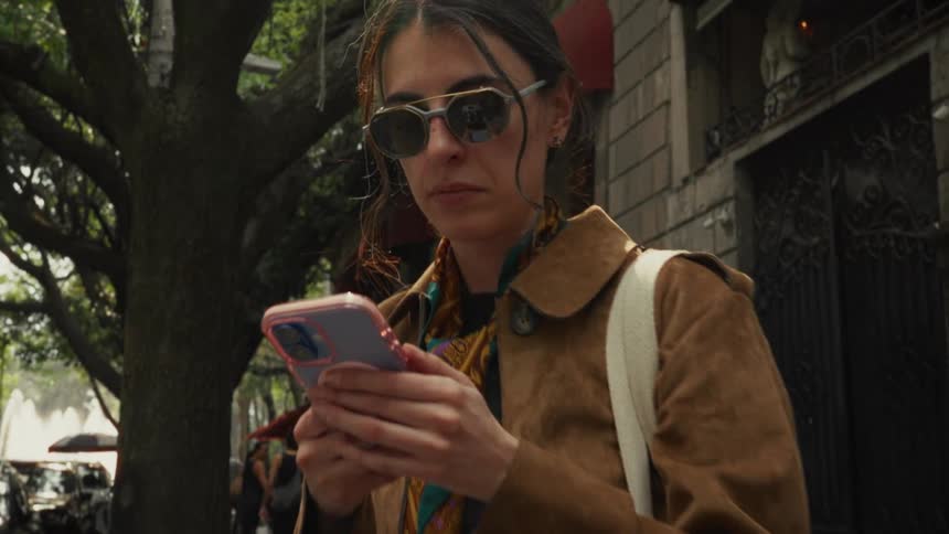 A stylish woman in sunglasses and a brown coat stands on a city street, intently focused on her phone