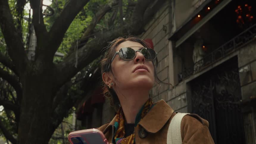 A stylish woman in sunglasses holds her phone while looking up at the architecture on a tree-lined city street