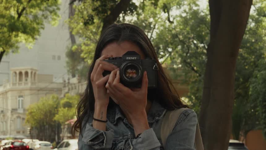 A young female photographer holds up a vintage film camera and takes a picture on a sunny city street