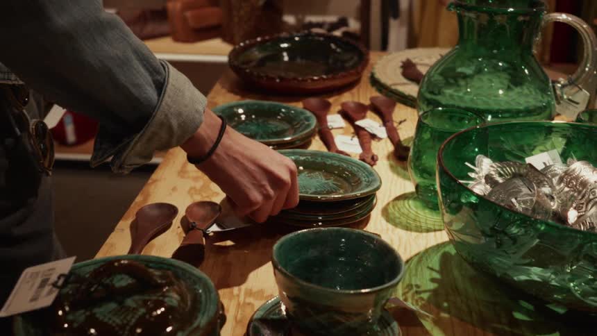 A person browses a collection of beautiful green handmade pottery and glassware on a wooden table in a boutique shop
