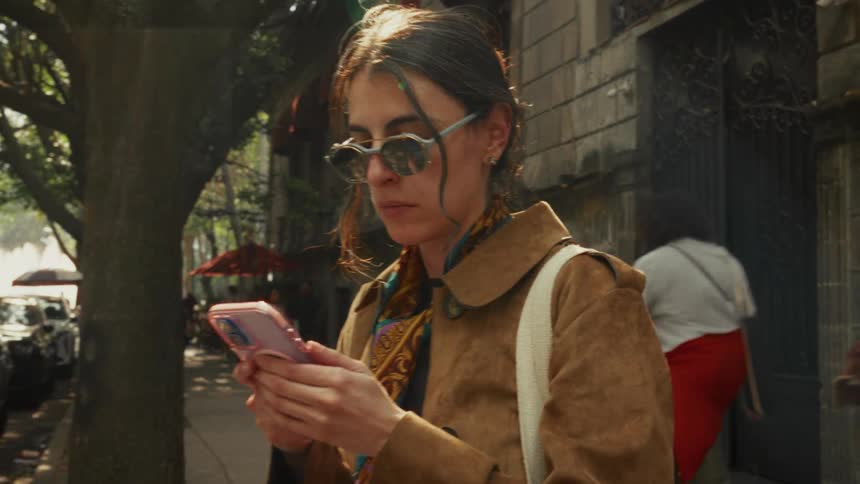 A stylish woman wearing sunglasses concentrates on her smartphone while standing on a sunny, tree-lined city street