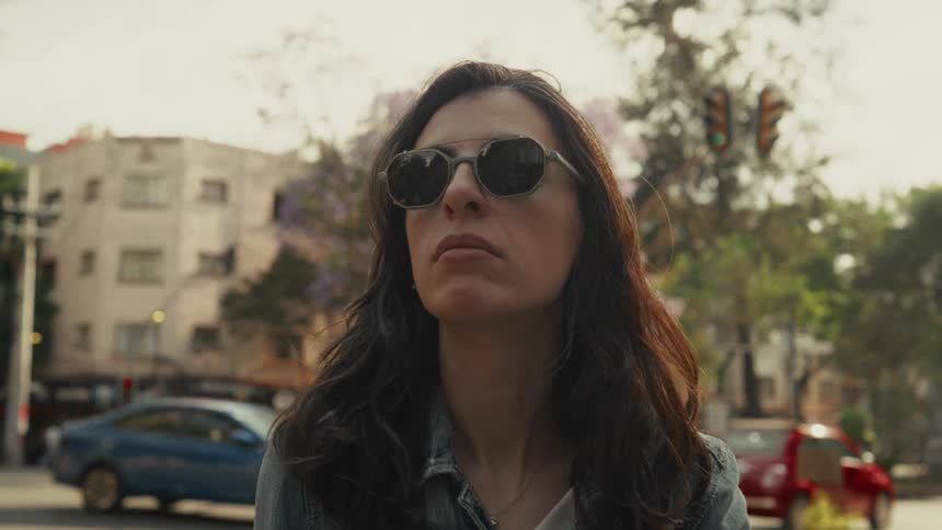 A contemplative woman in sunglasses stands on a bustling city street as the camera slowly zooms in on her face