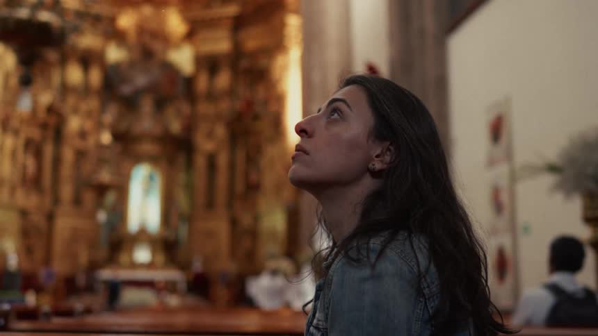 A pensive young woman looks up in reverence while sitting in a pew inside a beautiful, ornate church