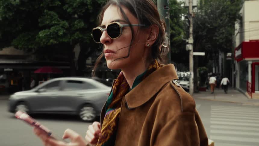 Stylish woman in sunglasses and a brown coat waits on a bustling city street corner while using her smartphone