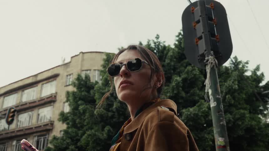 Cinematic low-angle shot of a pensive woman in sunglasses looking up while standing on a city street corner