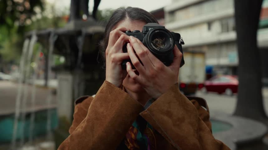 A female photographer raises a vintage film camera to take a picture while standing in a city park in front of a fountain