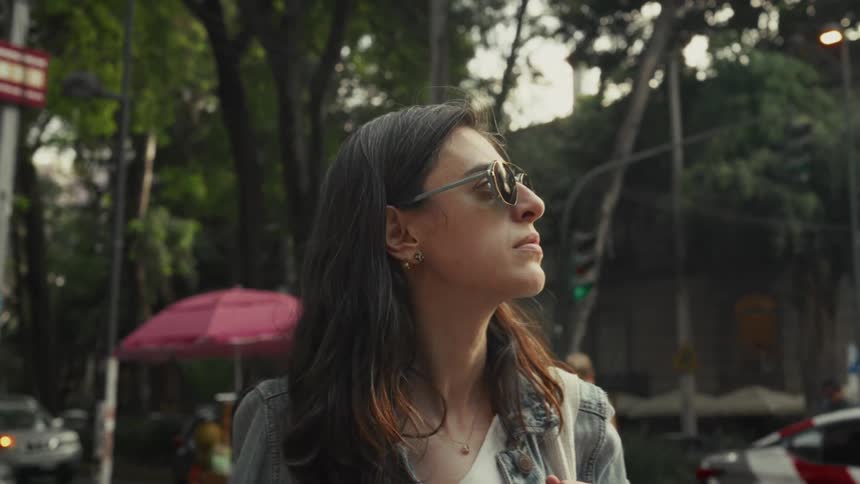 A thoughtful woman in sunglasses stands on a busy city street corner, observing the urban scene