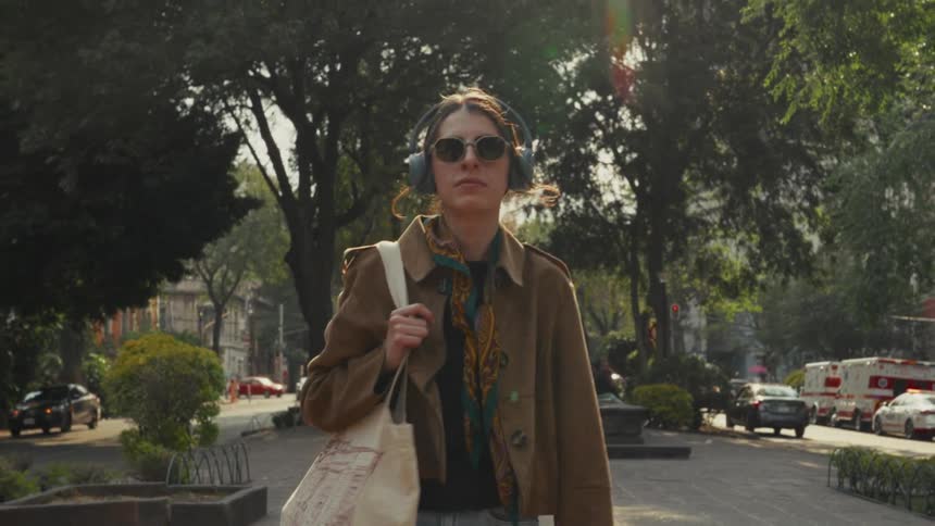 A stylish young woman wearing headphones and sunglasses walks towards the camera in slow motion through a sunny urban park