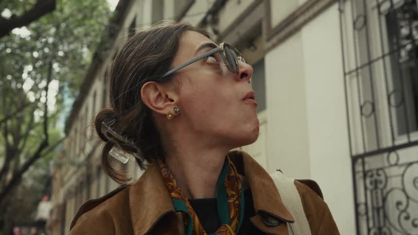 A woman in sunglasses enjoys a snack from a small glass dish while standing on a city street in slow motion