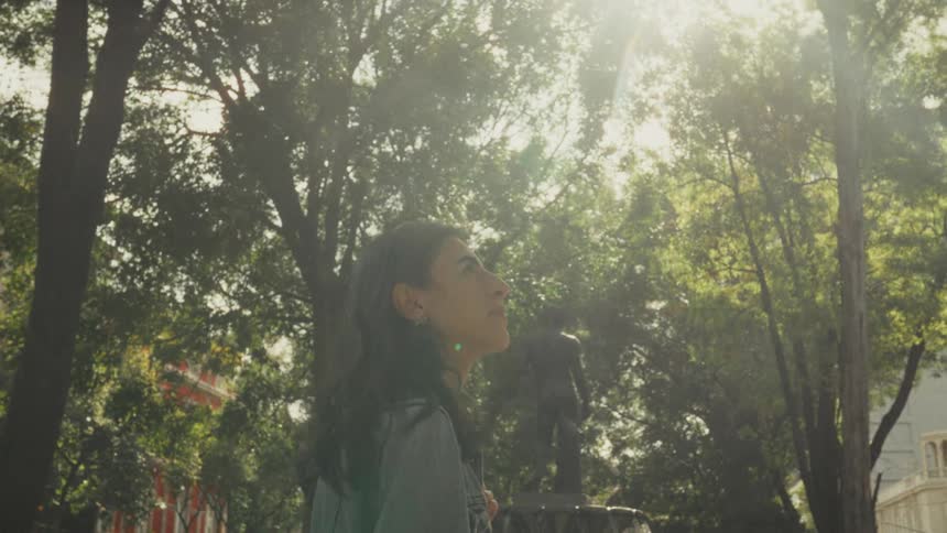 A thoughtful woman looks up at the sun shining through the trees in a beautiful city park