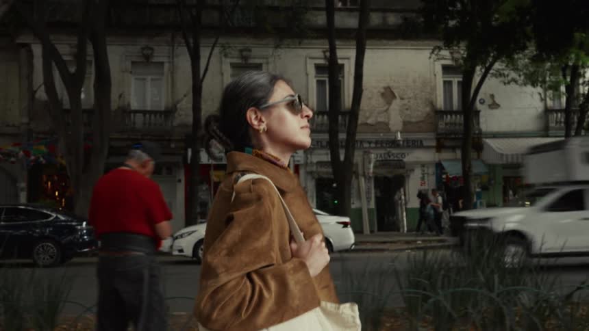 A stylish woman in sunglasses walks along a bustling city sidewalk in slow motion, with traffic and old buildings in the background
