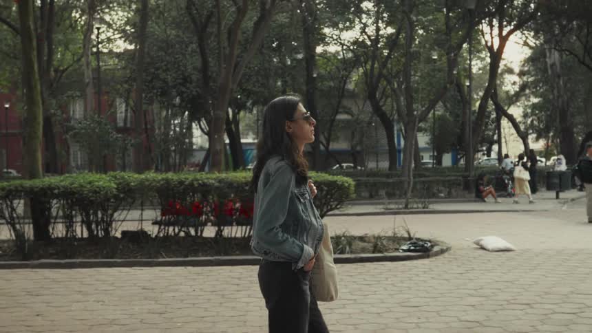 A stylish woman in a denim jacket and sunglasses walks casually through a lush city park in slow motion