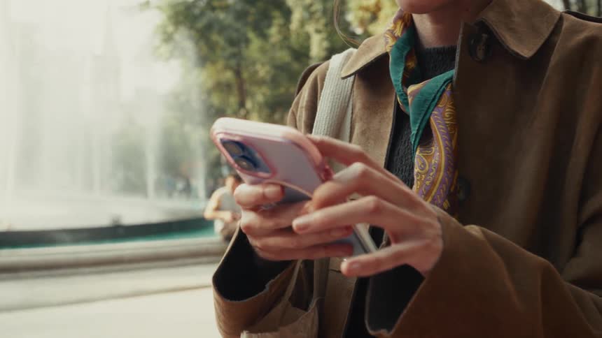 Close-up of a stylish woman's hands typing on a smartphone in a city park with a fountain in the background