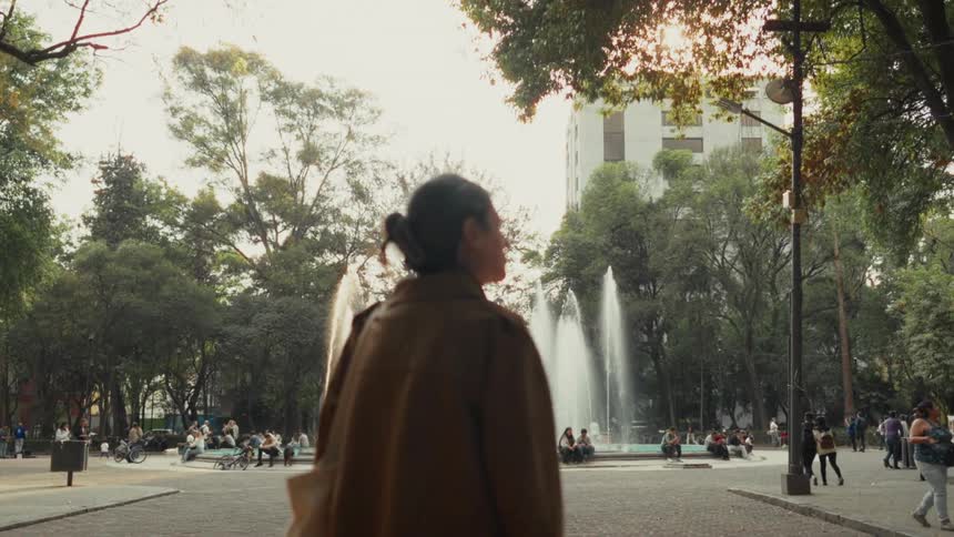 A woman from behind watches a water fountain in a busy urban park during golden hour