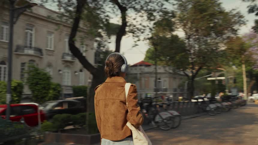A stylish woman wearing headphones walks along a sunny, tree-lined city street past moving traffic